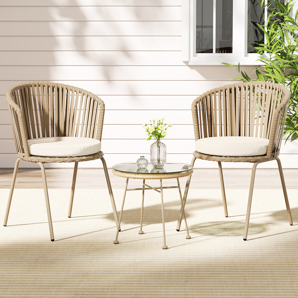 A three-piece outdoor bistro set including two beige chairs with rope weaving and a matching beige coffee table with a glass top, glass vase with flowers and bowl on the table, on a patio setting.