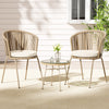 A three-piece outdoor bistro set including two beige chairs with rope weaving and a matching beige coffee table with a glass top, glass vase with flowers and bowl on the table, on a patio setting.