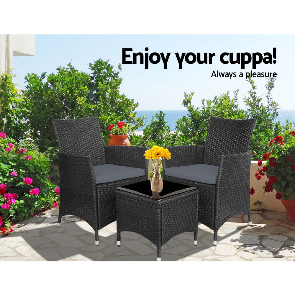 A three-piece outdoor bistro set consisting of two black wicker chairs with cushions and a matching black wicker side table with a glass top with a vase of flowers on it, set on a slate paved area in front of trees and flowers with the beach in the background.