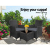 A three-piece outdoor bistro set consisting of two black wicker chairs with cushions and a matching black wicker side table with a glass top with a vase of flowers on it, set on a slate paved area in front of trees and flowers with the beach in the background.