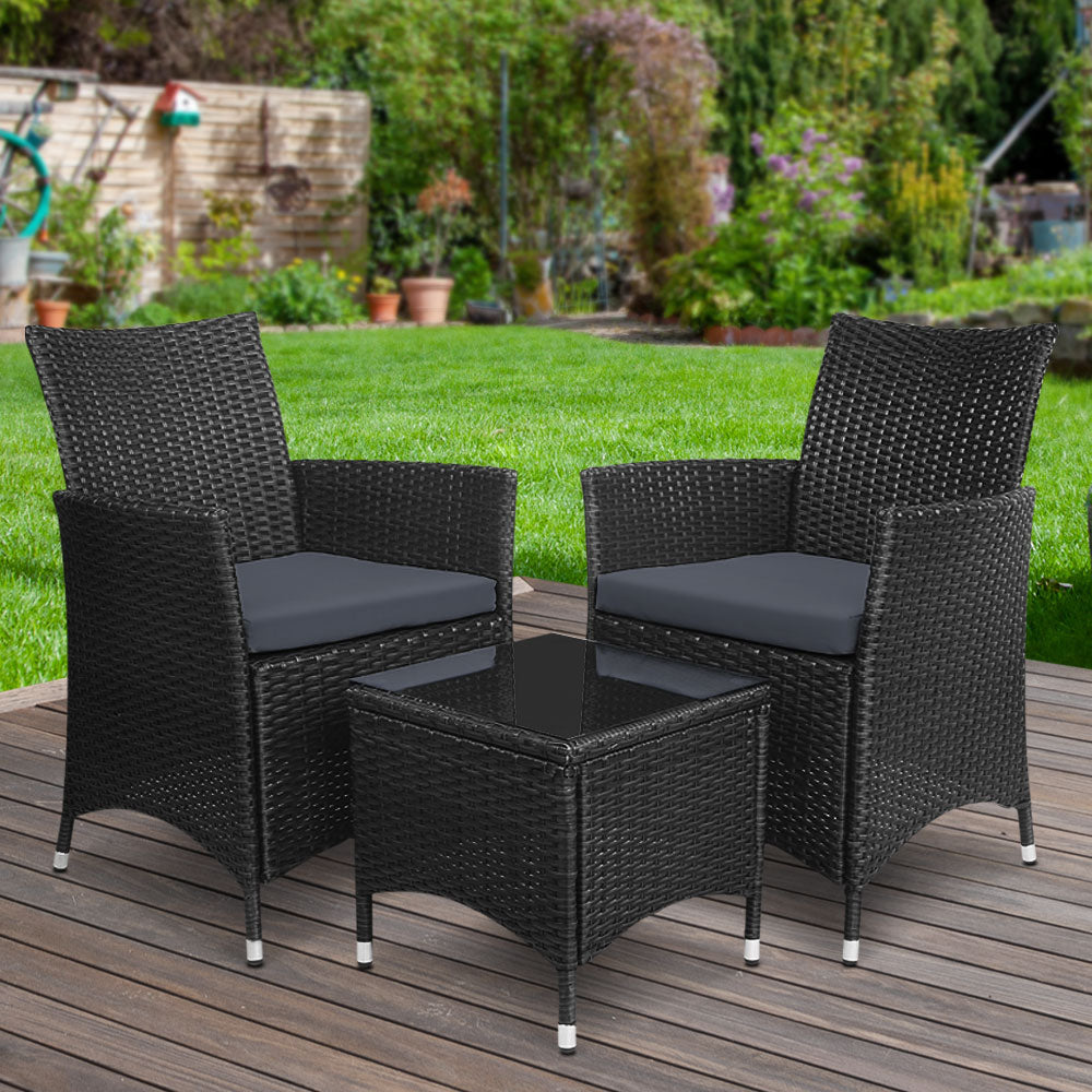 A three-piece outdoor bistro set consisting of two black wicker chairs with cushions and a matching black wicker side table with a glass top, on a wooden deck in a garden setting with green lawn, trees and flowers in the background.