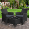A three-piece outdoor bistro set consisting of two black wicker chairs with cushions and a matching black wicker side table with a glass top, on a wooden deck in a garden setting with green lawn, trees and flowers in the background.