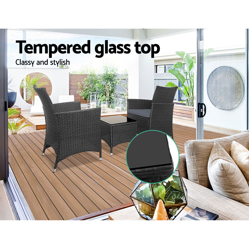 Three-piece outdoor bistro set with two black wicker chairs with cushions and a matching black wicker side table with glass top, on a wooden deck in a patio setting with water feature, plants and three candles in large glass holders in the backdrop.