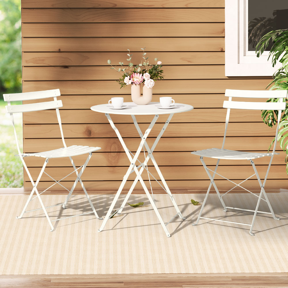 A Gardeon 3PC Outdoor Bistro Set consisting of a folding Table and Chairs in White, with 2 cups and saucers and a vase of flowers on the table, all set on a wood deck on a weaved and striped floor mat with a wooden house and garden in tthe background.