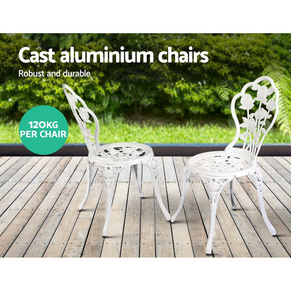 Two white chairs with rose design, set on a wooden decking in front of a grass and greenery in the backdrop.