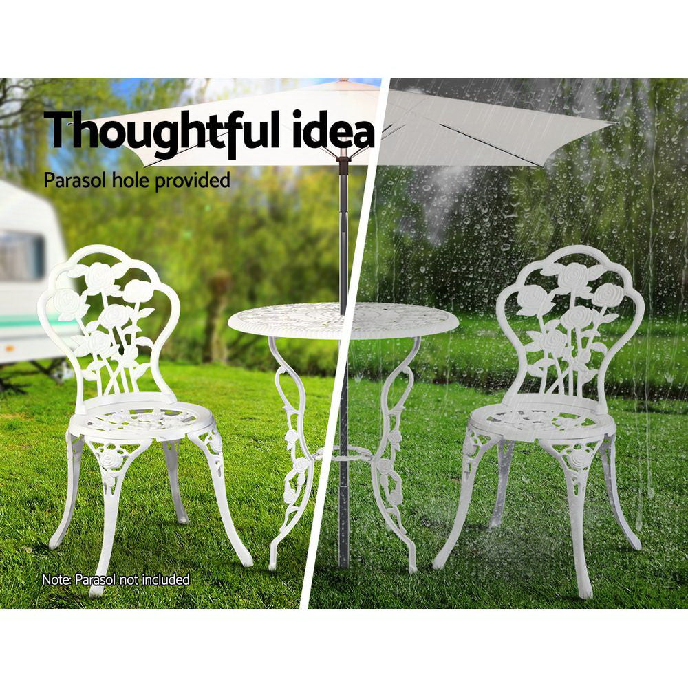 A white Gardeon bistro set including two chairs and a round table, made of cast aluminum with floral patterns, set on a grass and trees in the backdrop. The image is split into two showcasing the parasol hole holding a parasol in the middle of the table, half image in the sunshine offering shade and the other half in the rain also offering cover.