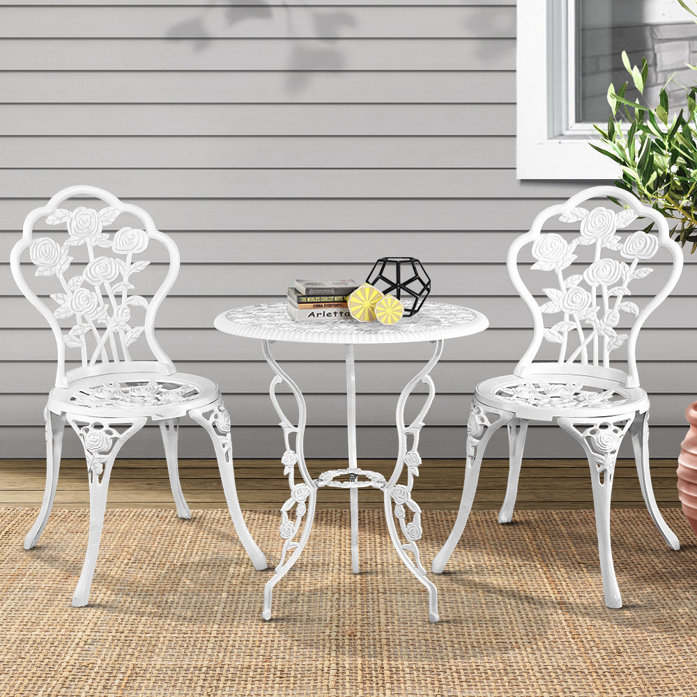 A white Gardeon bistro set including two chairs and a round table, with some books and ornaments on tabletop, made of cast aluminum with floral patterns, set in front of a grey wood house wall, set on a wooden deck and weaved floor mat, next to a planter with tree and window. 