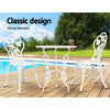 A white Gardeon bistro set including two chairs and a round table, made of cast aluminum with floral patterns, set on a wooden deck next to a pool with grass, trees and sky in the backdrop.