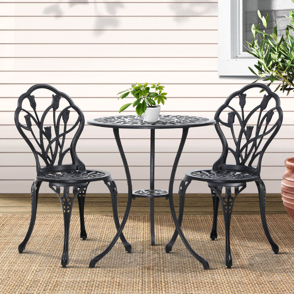 A black Gardeon 3-piece patio furniture set including a round table with potted plant on it, two chairs with intricate cut-out tulip patterns, set on a weaved floor mat in front of a wooden house wall with a window and plant to the side.