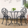 A black Gardeon 3-piece patio furniture set including a round table with potted plant on it, two chairs with intricate cut-out tulip patterns, set on a weaved floor mat in front of a wooden house wall with a window and plant to the side.