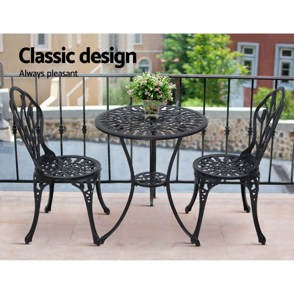 A black Gardeon 3-piece patio furniture set including a round table with an arrangement of flowers and two chairs with intricate cut-out tulip patterns set on a paved rooftop beside a metal fence with neighbouring terrace style houses in the backdrop.