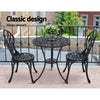 A black Gardeon 3-piece patio furniture set including a round table with an arrangement of flowers and two chairs with intricate cut-out tulip patterns set on a paved rooftop beside a metal fence with neighbouring terrace style houses in the backdrop.
