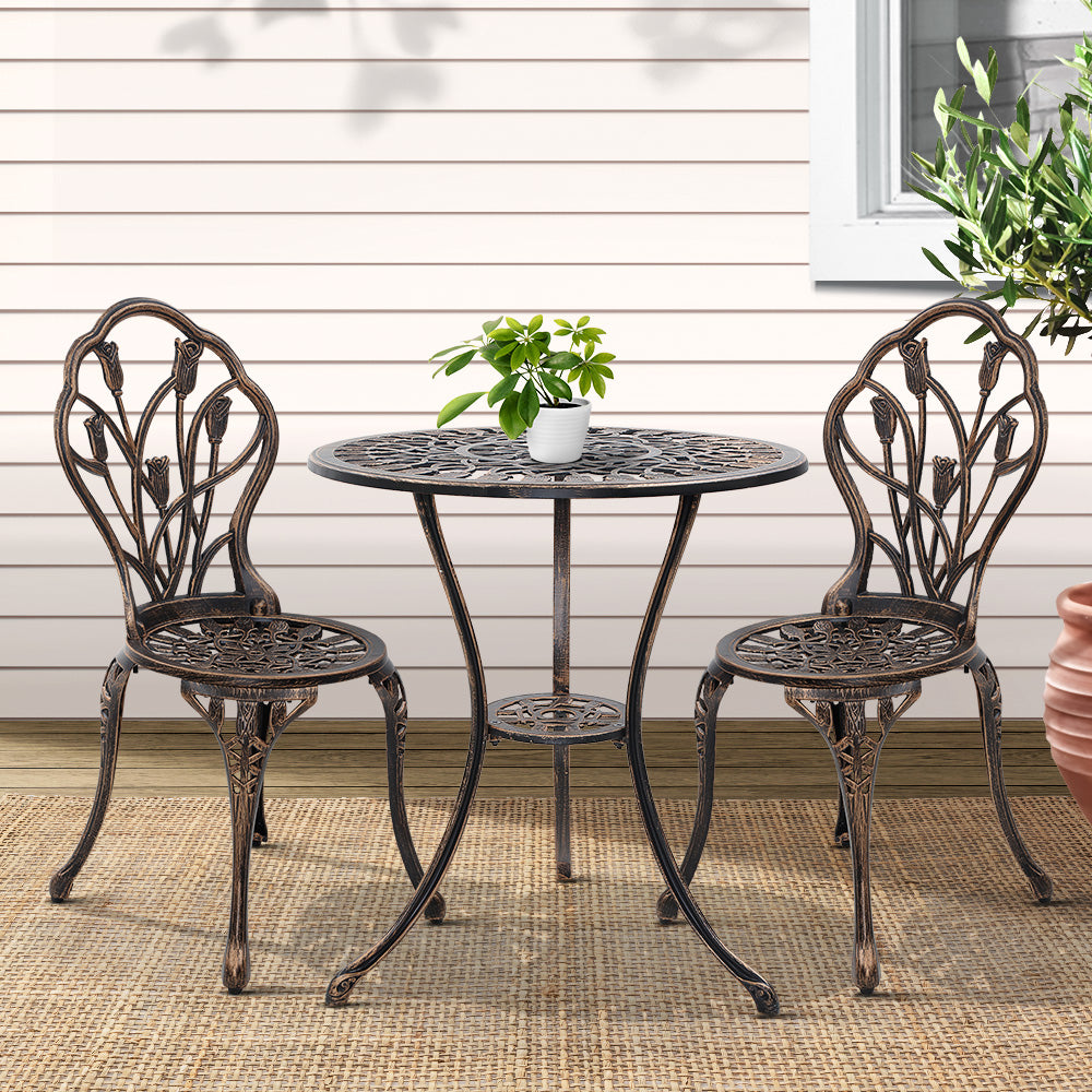 A bronze Gardeon 3-piece patio furniture set including a round table with potted plant on it, two chairs with intricate cut-out tulip patterns, set on a weaved floor mat in front of a wooden house wall with a window and plant to the side.