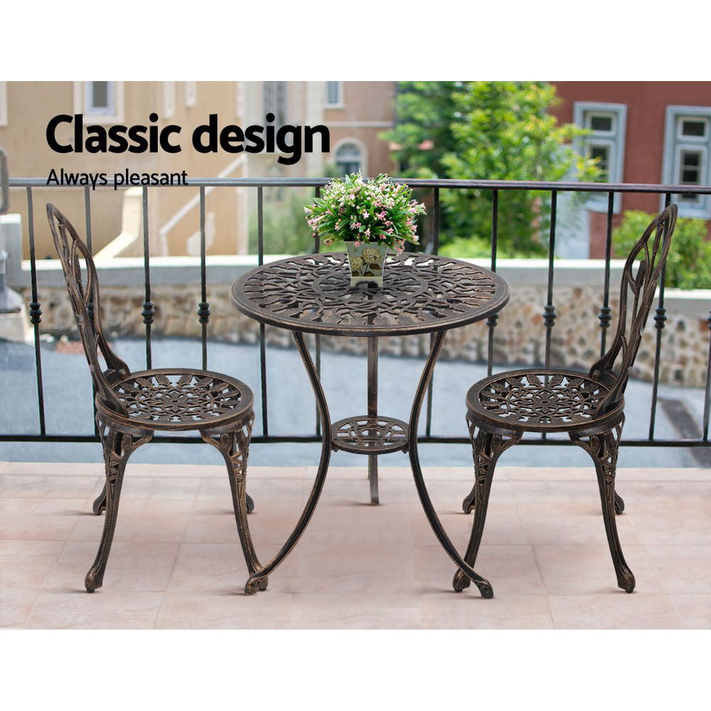 A bronze Gardeon 3-piece patio furniture set including a round table with an arrangement of flowers and two chairs with intricate cut-out tulip patterns set on a paved rooftop beside a metal fence with neighbouring terrace style houses in the backdrop.