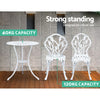 A white Gardeon bistro set including two chairs and a round table, made of cast aluminum with tulip patterns, set on a laminated wood floor before a grey tiled wall, one chair facing forward and one facing backwards, shows the weight capacity being 40kg for the table and 120kg for the chairs.