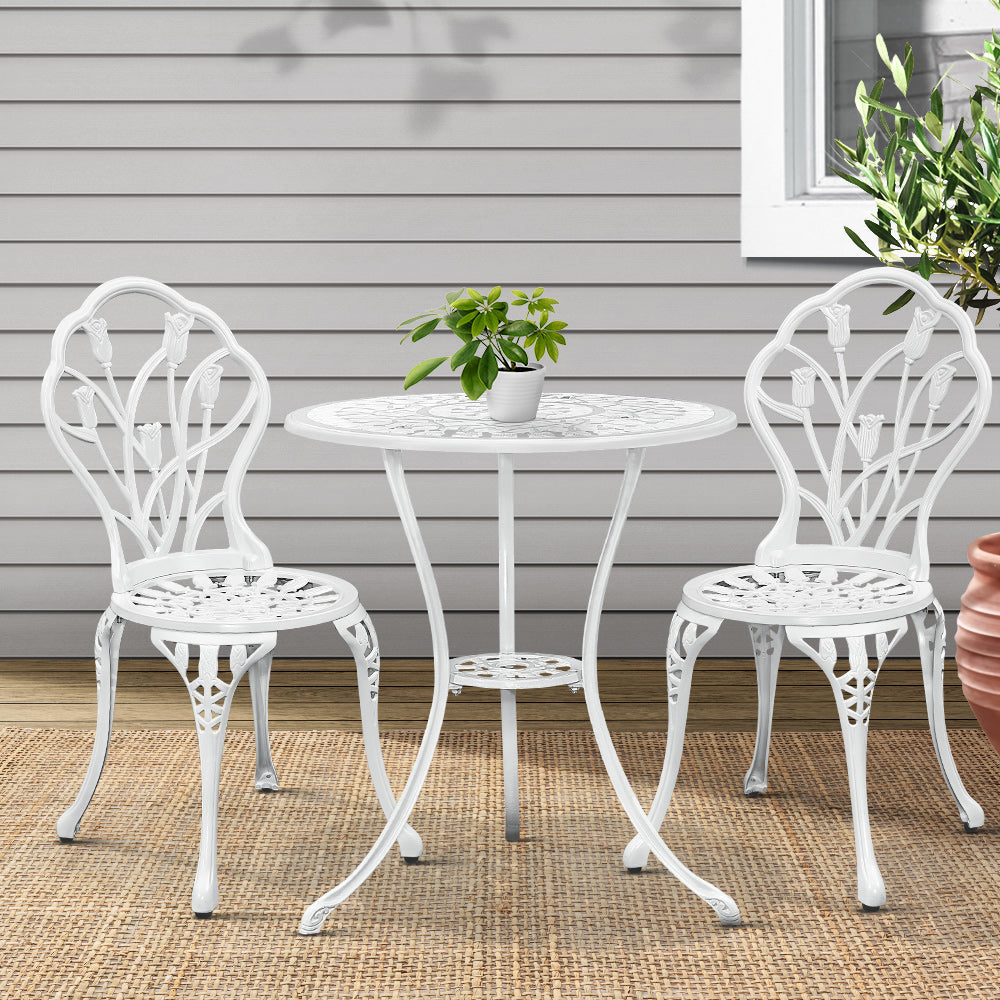A white Gardeon 3-piece patio furniture set including a round table with potted plant on it, two chairs with intricate cut-out tulip patterns, set on a weaved floor mat in front of a wooden house wall with a window and plant to the side.