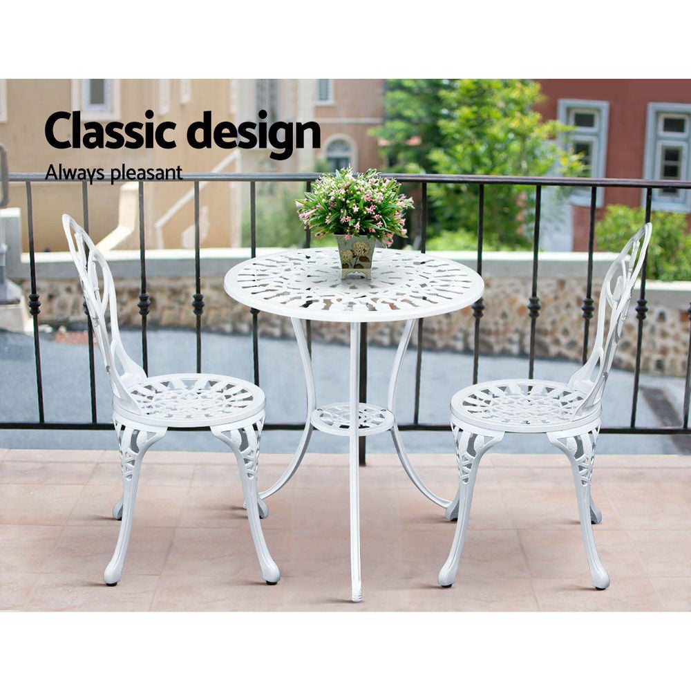 A white Gardeon 3-piece patio furniture set including a round table with an arrangement of flowers and two chairs with intricate cut-out tulip patterns set on a paved rooftop beside a metal fence with neighbouring terrace style houses in the backdrop.