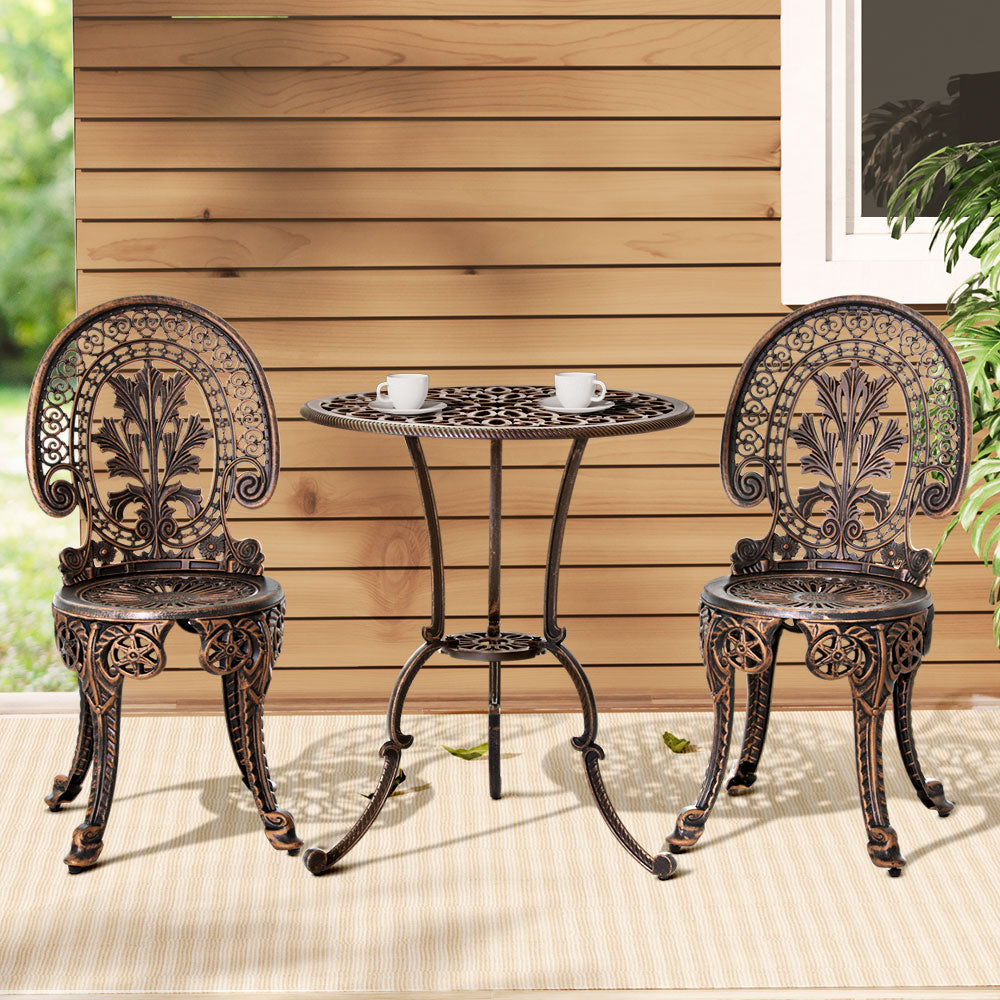 A bronze Gardeon 3-piece patio furniture set including a round table with 2 cups and saucers on it, two chairs with intricate cut-out patterns, set on a white and beige striped floor mat in front of a wooden house wall with a window and plant to the side.