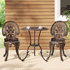 A bronze Gardeon 3-piece patio furniture set including a round table with 2 cups and saucers on it, two chairs with intricate cut-out patterns, set on a white and beige striped floor mat in front of a wooden house wall with a window and plant to the side.
