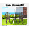 A bronze Gardeon bistro set including two chairs and a round table, made of cast aluminum with inticate designs, set on a grass with hedging and an ocean view in the backdrop. The image is split into two showcasing the parasol hole holding a parasol in the middle of the table, half image in the sunshine offering shade and the other half in the rain also offering cover.