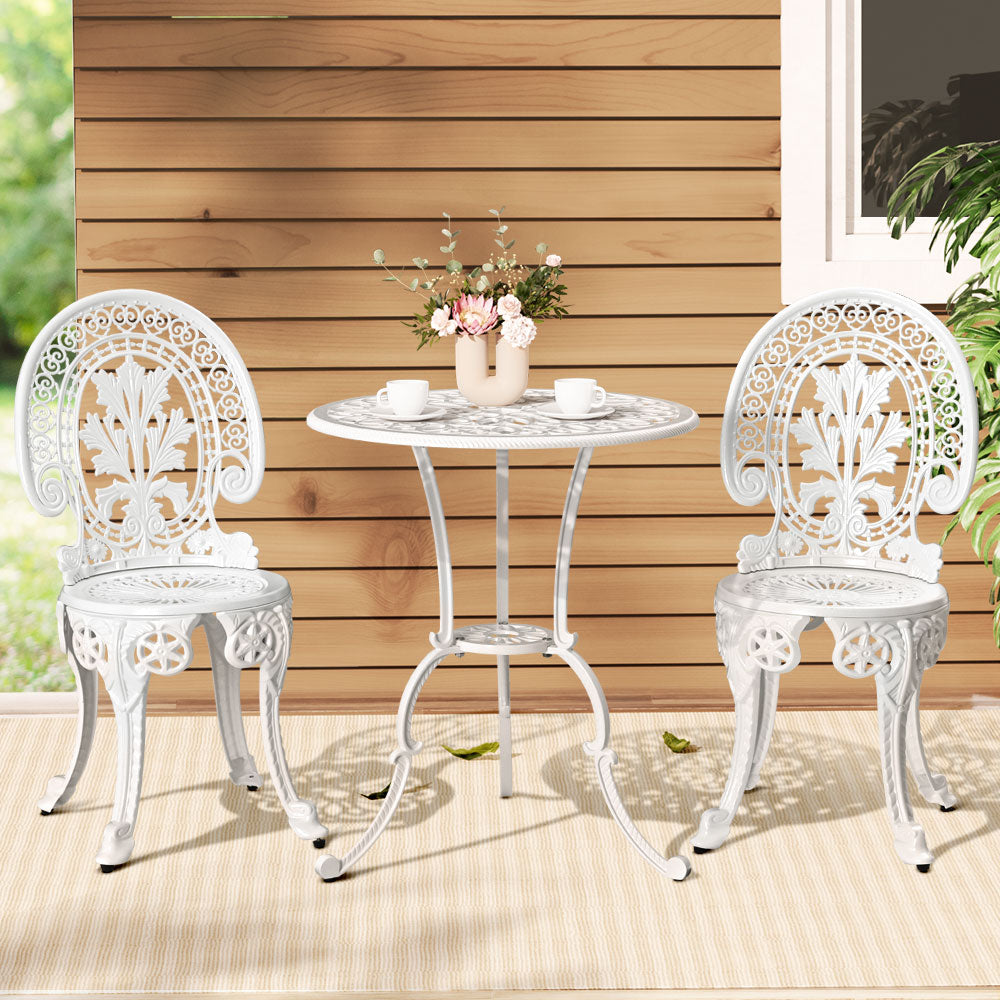 A white Gardeon 3-piece patio furniture set including a round table with 2 cups and saucers and a vase of flowers on it, two chairs with intricate cut-out patterns, set on a white and beige striped floor mat in front of a wooden house wall with a window and plant to the side.