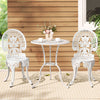 A white Gardeon 3-piece patio furniture set including a round table with 2 cups and saucers and a vase of flowers on it, two chairs with intricate cut-out patterns, set on a white and beige striped floor mat in front of a wooden house wall with a window and plant to the side.