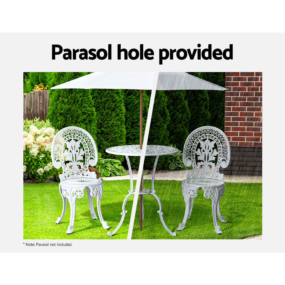 A white Gardeon bistro set including two chairs and a round table, made of cast aluminum with inticate designs, set on a grass and trees and flowers in the backdrop. The image is split into two showcasing the parasol hole holding a parasol in the middle of the table, half image in the sunshine offering shade and the other half in the rain also offering cover.