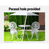 A white Gardeon bistro set including two chairs and a round table, made of cast aluminum with inticate designs, set on a grass and trees and flowers in the backdrop. The image is split into two showcasing the parasol hole holding a parasol in the middle of the table, half image in the sunshine offering shade and the other half in the rain also offering cover.