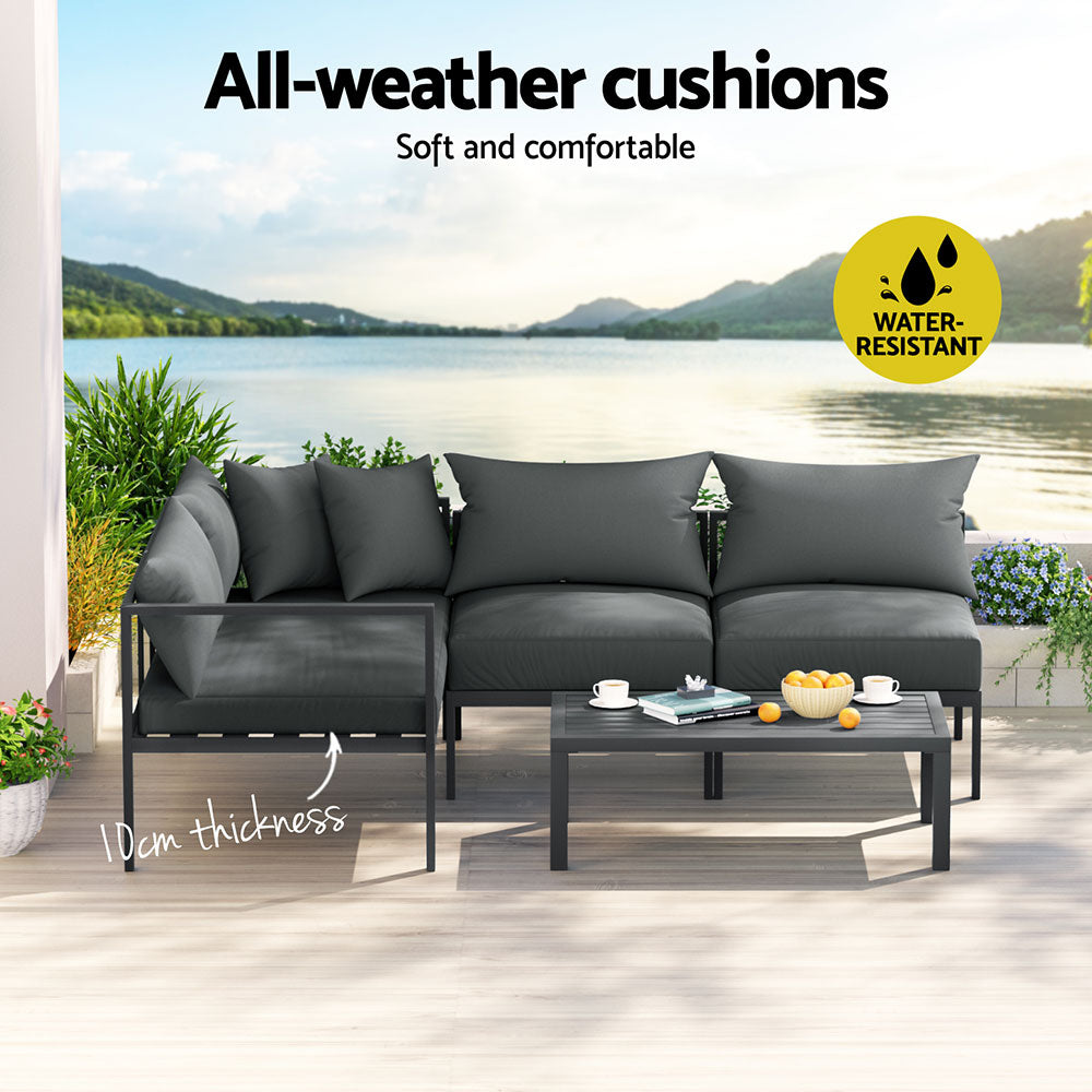 A Gardeon 4-seater Outdoor Aluminium Sofa Set with a two-seater sofa, two single sofas and a wide coffee table in charcoal black colour, seats are together forming an 'L' shape and set on a decked outdoor area with a lake view in the background.
