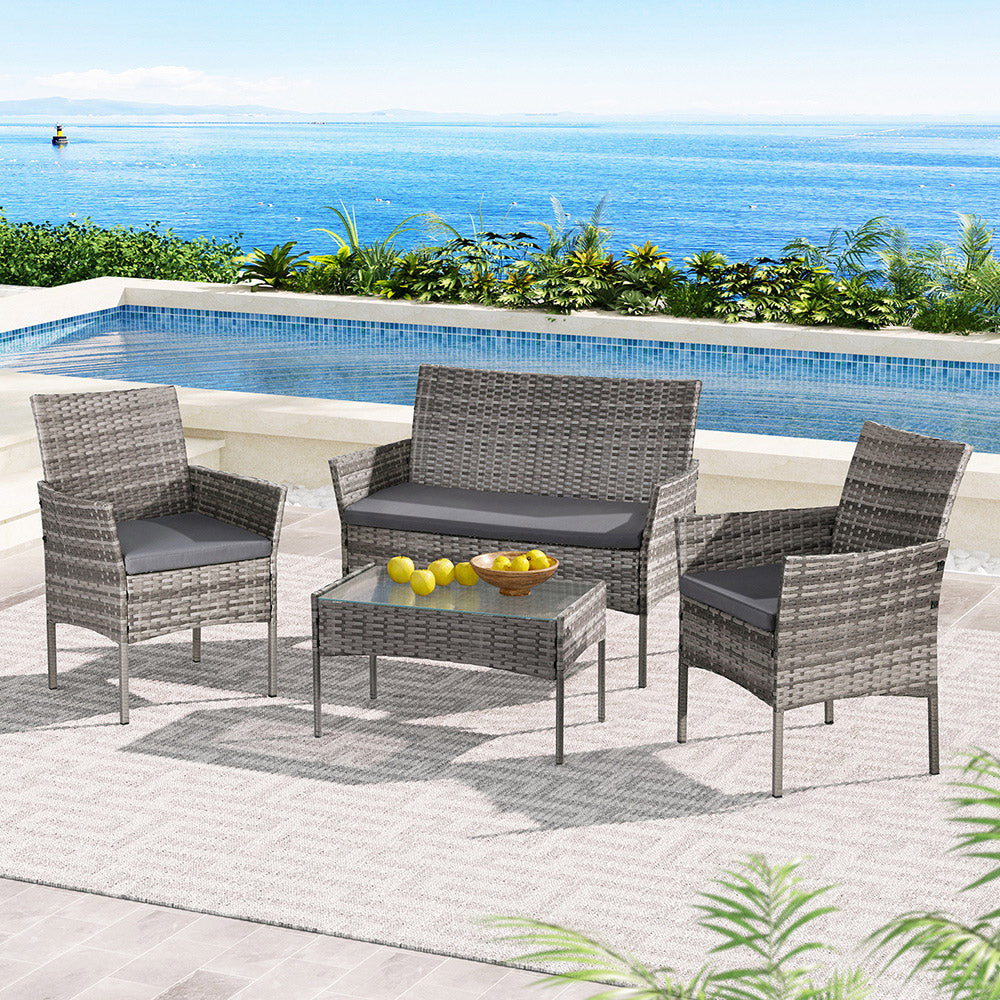 Gardeon 4-Seater Outdoor Wicker Sofa Set featuring a glass top Table with bowl of lemons on it, a two-seater and two single Chairs in Grey. All set on a poolside balcony with plants and an ocean view in the background.