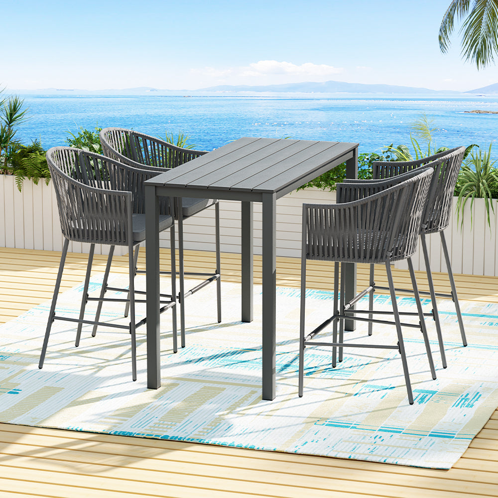 Five-piece outdoor bar set with a rectangular dining table and four grey rope chairs with slanted backrests and handrails. Set on a patterned floor mat on a wooden deck in front of wooden planters and plants with an ocean view in the backdrop.