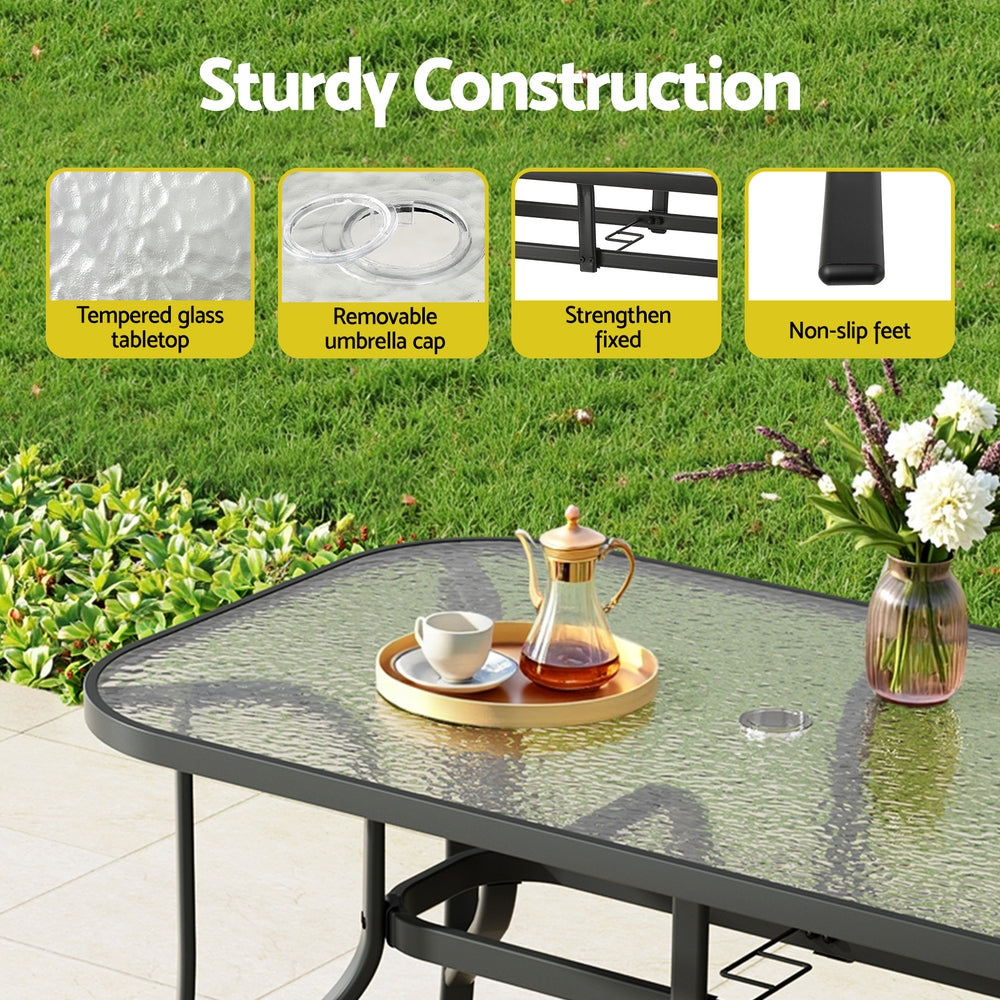 Outdoor table with tempered glass top, removable umbrella cap, strengthened fixed parts, and non-slip feet on a grassy background.