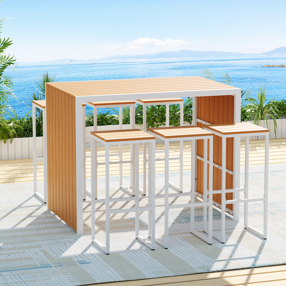 Seven-piece bar set with a rectangular table and six bar stools with a wooden slat tabletop and white steel frames. Set on a patterned floor mat on a wooden deck in front of wooden planters and plants with an ocean view in the backdrop.