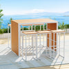 Seven-piece bar set with a rectangular table and six bar stools with a wooden slat tabletop and white steel frames. Set on a patterned floor mat on a wooden deck in front of wooden planters and plants with an ocean view in the backdrop.