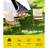 Two Gardeon portable folding beach chairs made of steel with beige fabric seats, set on grass in front of a tent with a small folding wooden table that has 2 cups and saucers on and a bowl of fruits.