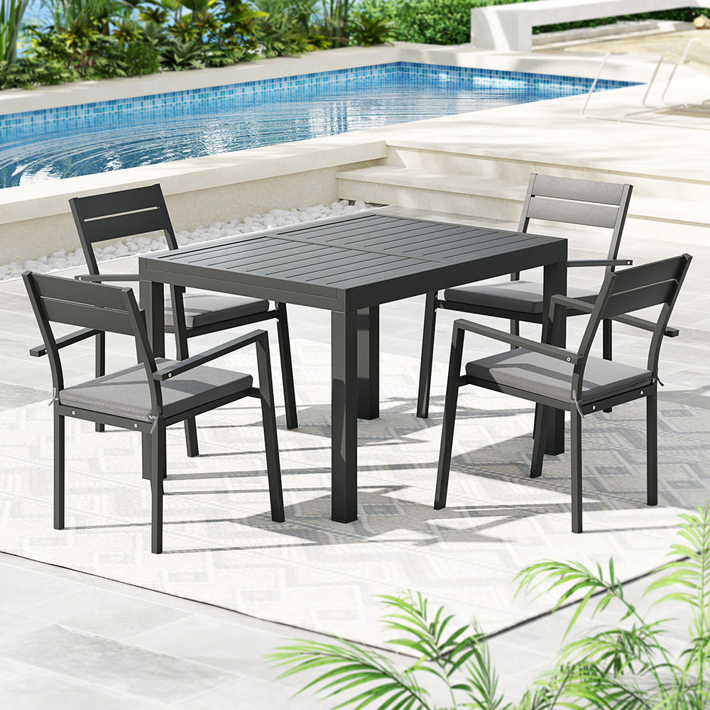 A five-piece outdoor dining set with a rectangular dining table and four grey aluminium chairs with cushions, all set at a poolside on a tiled flooring with a floor mat.