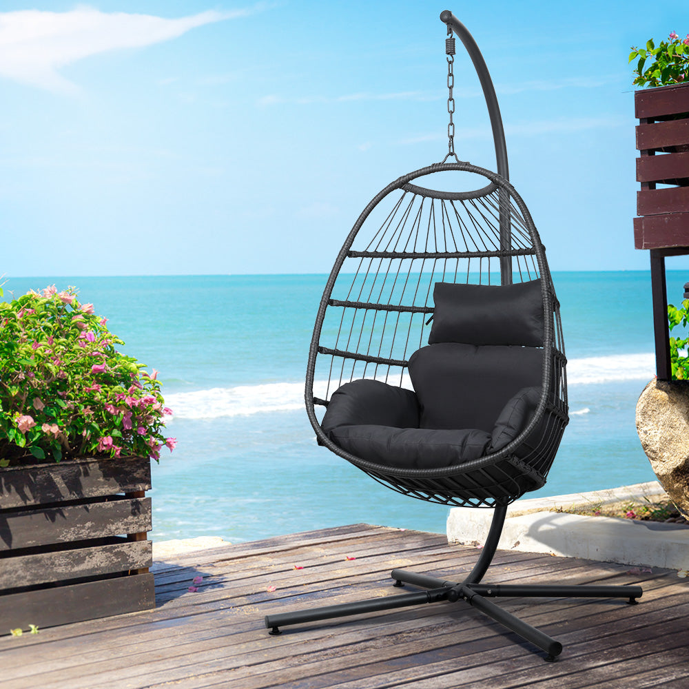 Hanging egg chair with black cushions on a wooden deck by the ocean.