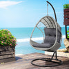 An outdoor egg swing chair with a grey wicker frame, charcoal stand and cushions on a wood deck beside the sea.