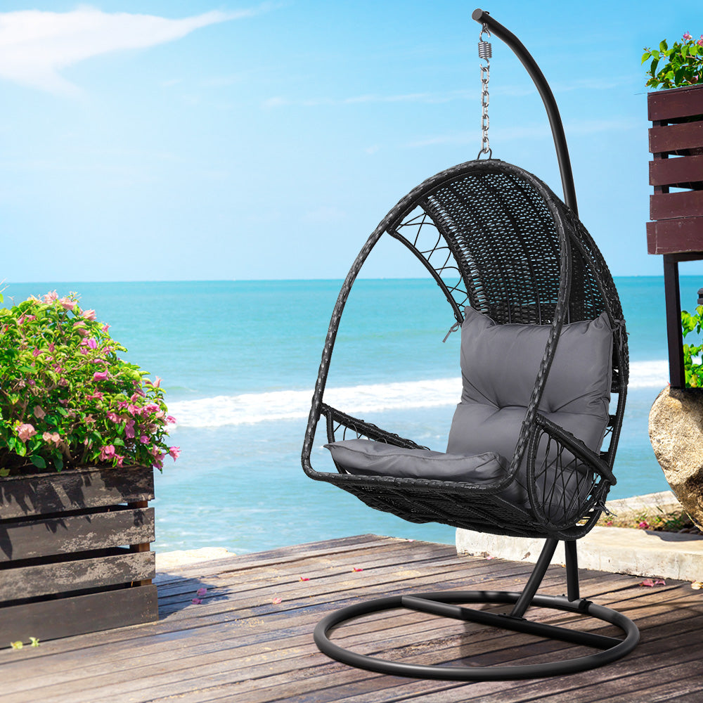 An outdoor egg swing chair with a black wicker frame and stand, featuring a grey cushion. Set on a wooden deck with ocean view in the background.