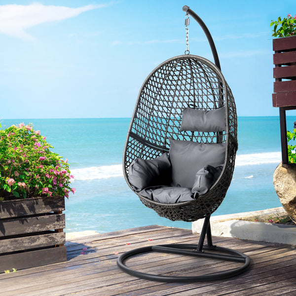 Woven egg-shaped hanging chair with gray cushions on a wooden deck by the ocean.