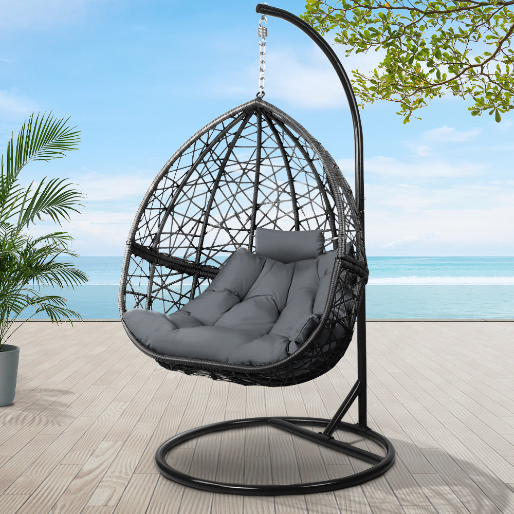 Hanging egg chair with gray cushion on a stand outdoors with a beach view.