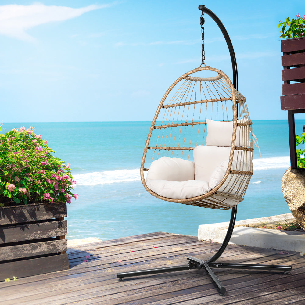 Wicker hanging chair with white cushions on a wooden deck by the ocean.