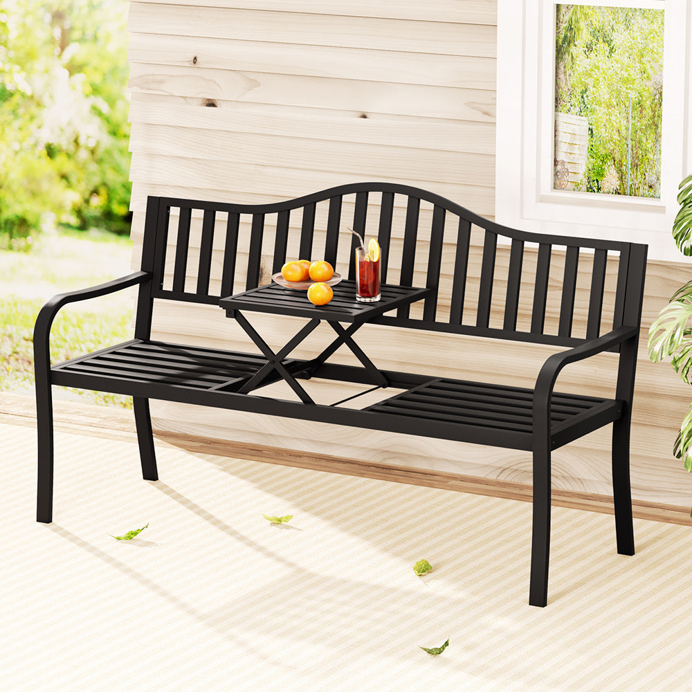 A black steel outdoor garden bench with a foldable table, featuring a slatted design for the seat and backrest, a glass of iced tea and a bowl of fruit on table, set on a patio area in front of a wooden house wall and window with garden in the background.