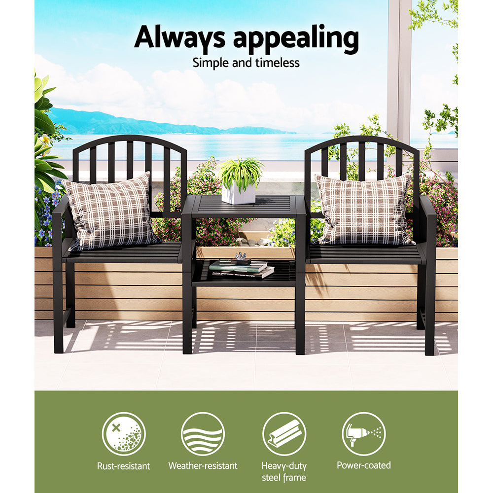 A two-seater garden bench set with a black steel frame, featuring an all-in-one table and armchairs, cushions on the seats, a plant pot on the table and books on the shelf under the table, set in front of a window overlooking an ocean view.