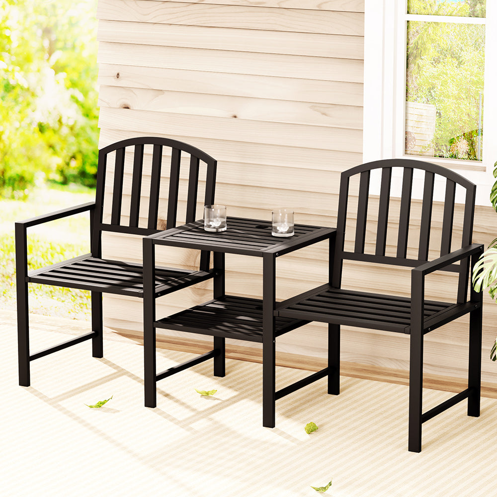 A two-seater garden bench set with a black steel frame, featuring an all-in-one table and armchairs, a couple of glasses with water on the table, set on a patio area in front of a wooden house wall and window with garden in the background.