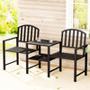 A two-seater garden bench set with a black steel frame, featuring an all-in-one table and armchairs, a couple of glasses with water on the table, set on a patio area in front of a wooden house wall and window with garden in the background.