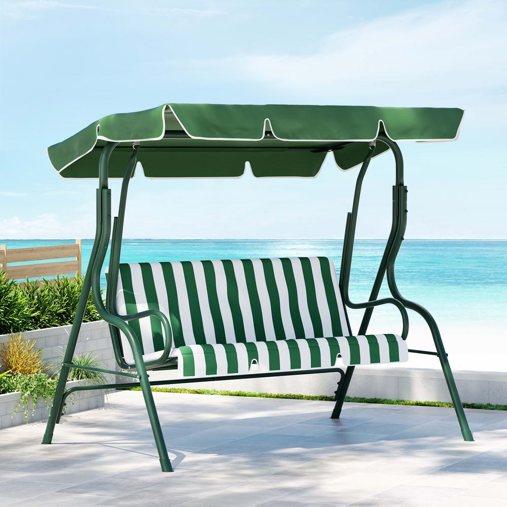 A white and green Gardeon outdoor swing chair with a canopy and striped cushions, in an outdoor patio setting overlooking the ocean.