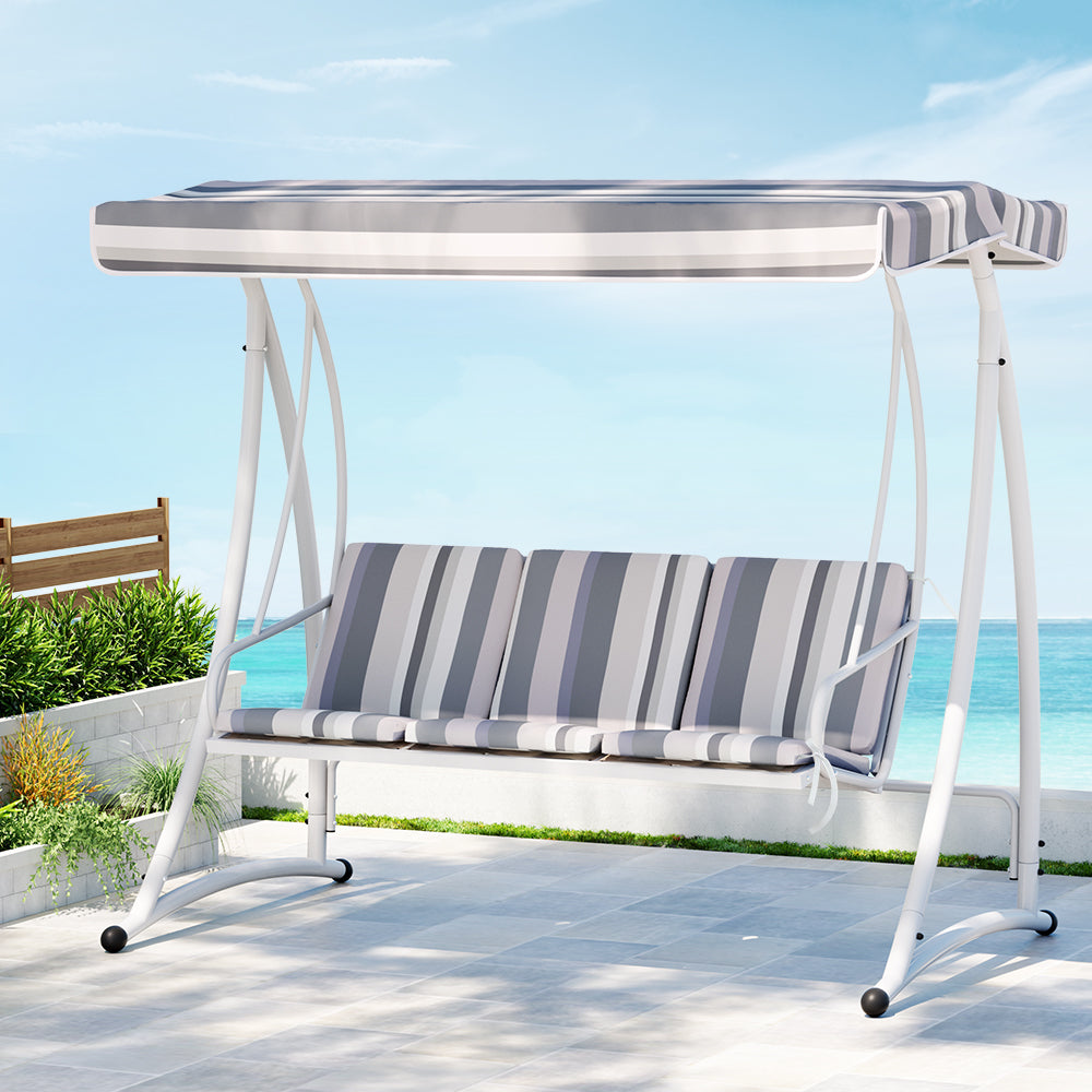 An outdoor swing chair with a white and grey striped canopy and a sturdy white frame, designed to seat three people and set on a tiled area with plants and an ocean view in the background.