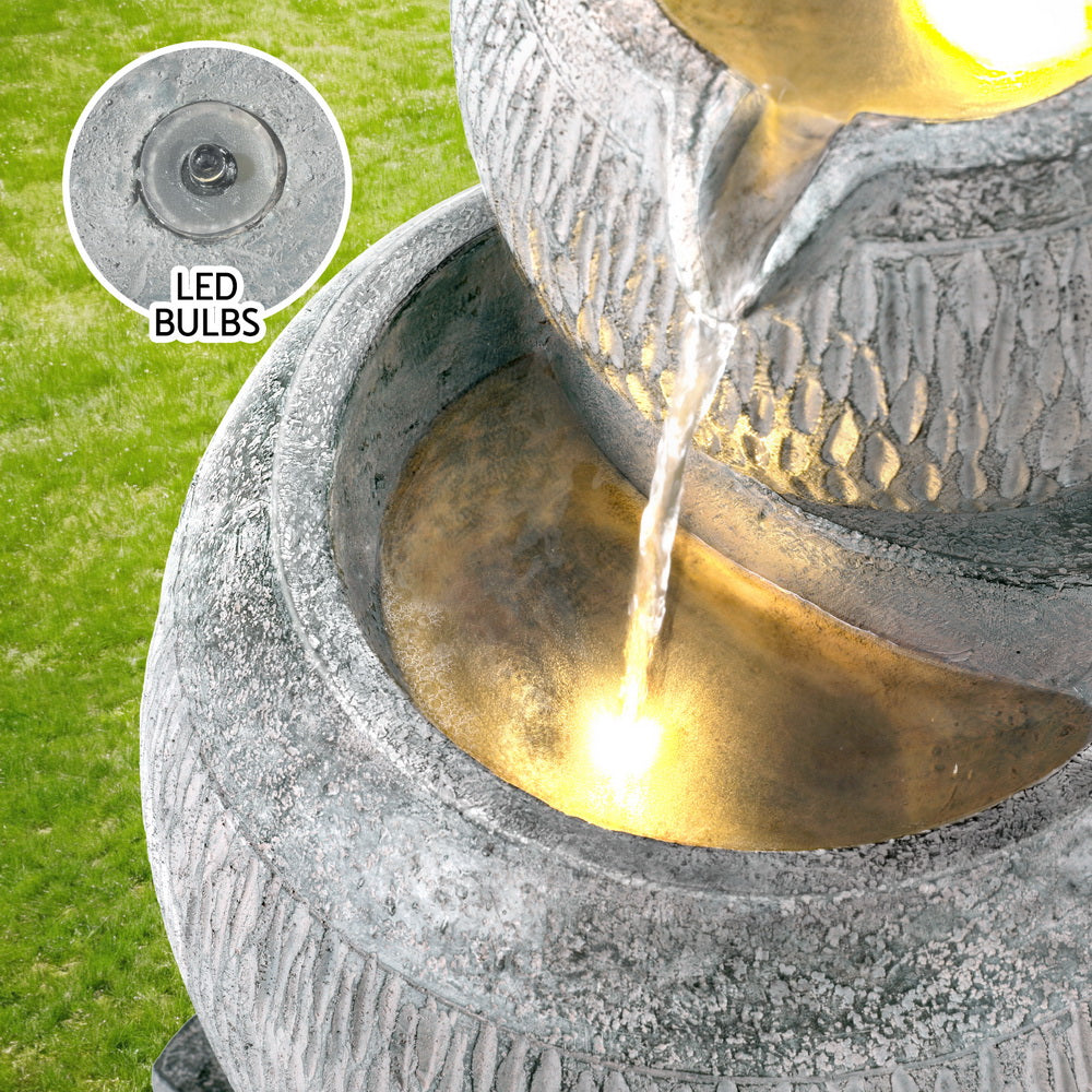 A close-up view of top bowl pouring into middle bowl of a three-tier cascading water fountain feature with LED lights, made of polyresin, in a grey colour, insert image of the LED bulb included, with green grass in the backdrop.