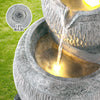 A close-up view of top bowl pouring into middle bowl of a three-tier cascading water fountain feature with LED lights, made of polyresin, in a grey colour, insert image of the LED bulb included, with green grass in the backdrop.